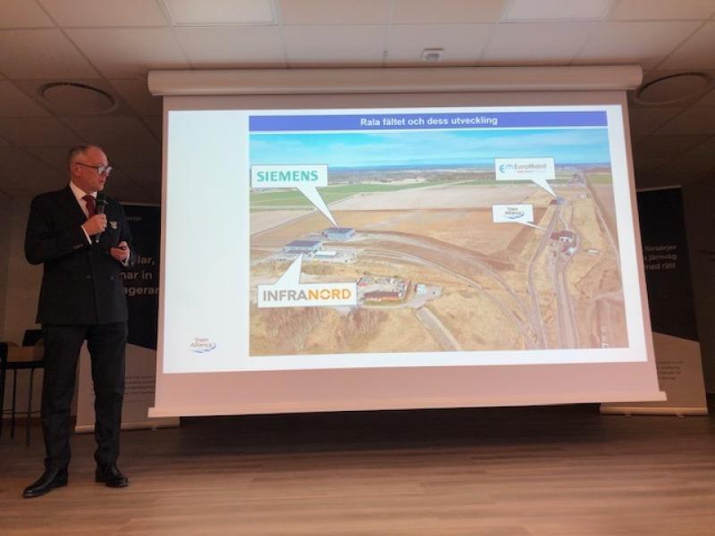 En kostymklädd man talar i en mikrofon bredvid en projicerad bild som visar en flygbild över ett industriområde med Siemens, Infranord och EnTurMobil.