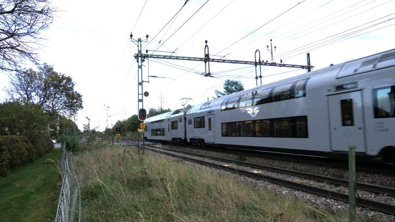 Ett silverfärgat dubbeldäckat passagerartåg färdas längs järnvägsspår under elektriska kontaktledningar och passerar en gräsyta och ett rött signalljus.