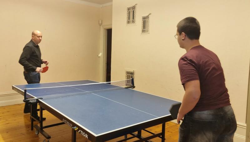 Två personer spelar bordtennis i ett inomhusrum med beige väggar och trägolv.