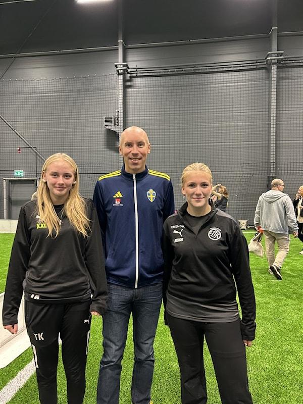 Tre personer står på ett konstgräs inomhus. Mannen i mitten bär en blå Sweden-jacka, de två flickorna på vardera sidan bär svarta sportkläder. Andra personer syns i bakgrunden.