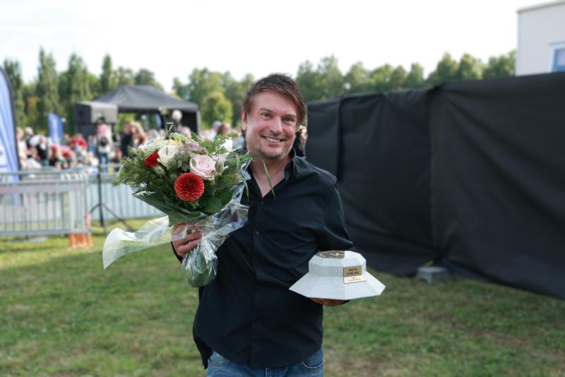 En man står utomhus med en blomsterbukett i ena handen och en pokal i den andra, och ler mot kameran.
