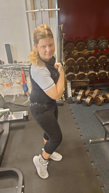 En kvinna poserar på ett gym och spänner armen bredvid ett ställ med hantlar. Hon har på sig ett grått och svart träningsset med vita skor.
