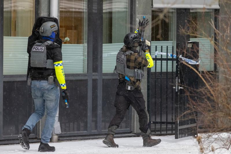 Två poliser i taktisk utrustning går in på ett inhägnat område utanför en byggnad en snöig dag, medan en av poliserna håller upp en polisavspärrning.