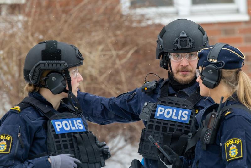 Utanför står tre svenska poliser i taktisk utrustning och pratar, iklädda hjälmar och västar med texten 