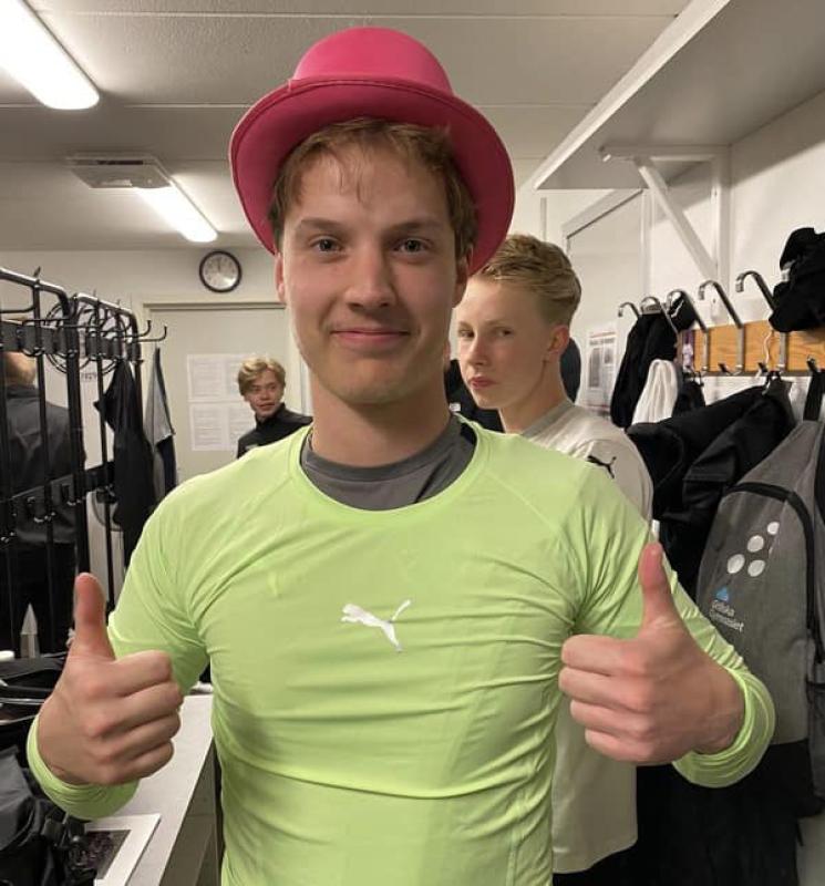 Man i limegrön skjorta och rosa hatt ger tummen upp i ett omklädningsrum med andra människor i bakgrunden.