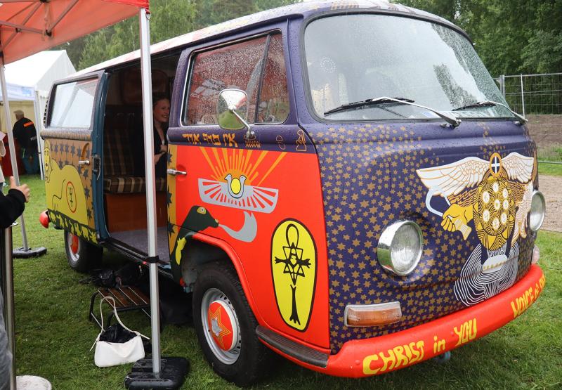 En gammal Volkswagen-skåpbil med färgglada, intrikata konstverk och symboler på utsidan är parkerad på gräset under ett skärmtak en regnig dag.