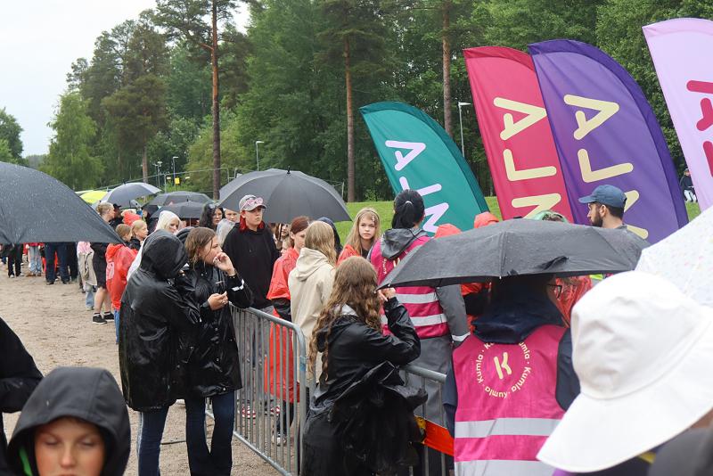 En folkmassa, många med paraplyer och regnrockar, väntar i kö bredvid färgglada flaggor och volontärer i rosa västar en regnig dag.