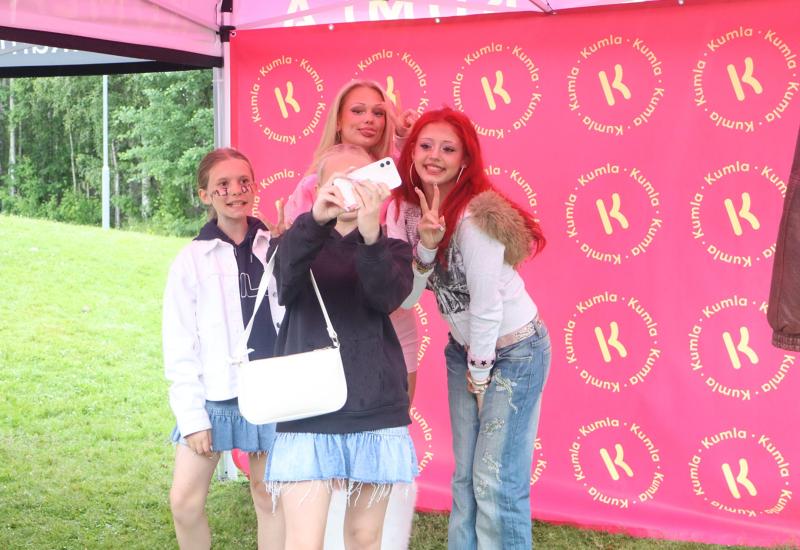 Fyra ungdomar poserar för en selfie framför en rosa bakgrund med Kumlas varumärke vid ett utomhusevenemang. Gräs och träd syns i bakgrunden.