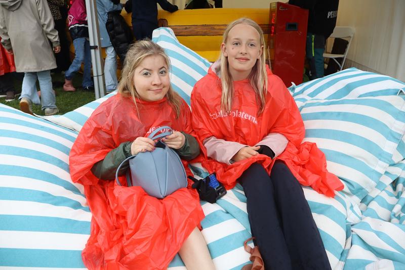 Två flickor i röda regnponchos sitter på blå- och vitrandiga sittsäckar utomhus, med andra människor och tält synliga i bakgrunden.