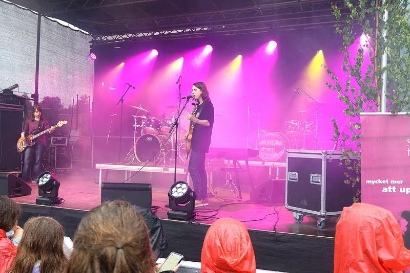 Ett band uppträder på en utomhusscen med lila belysning, sett av en publik i regnrockar en regnig dag.
