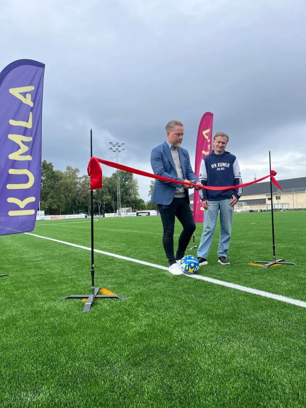 Två män står på en fotbollsplan bredvid ett rött band; den ene mannen håller i en sax och en fotboll och förbereder sig för en bandklippningsceremoni. Lila banderoller med texten ”KUMLA” syns.