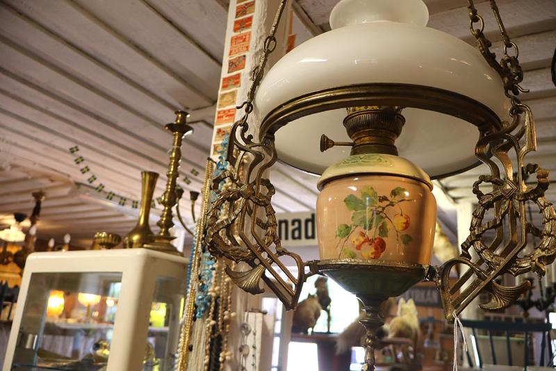 En vintage-oljelampa att hänga upp, med blommönster och detaljer i mässing, omgiven av antikviteter, däribland ljusstakar i mässing och ett glasvitrinskåp, i en butik.