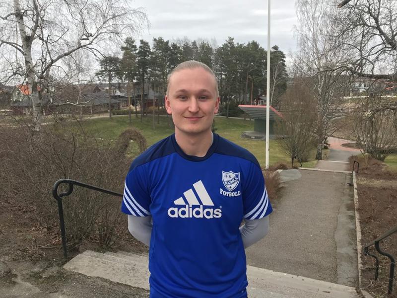 En person i en blå Adidas fotbollströja står utomhus på en betongtrappa, med lövlösa träd och ett parkområde i bakgrunden.