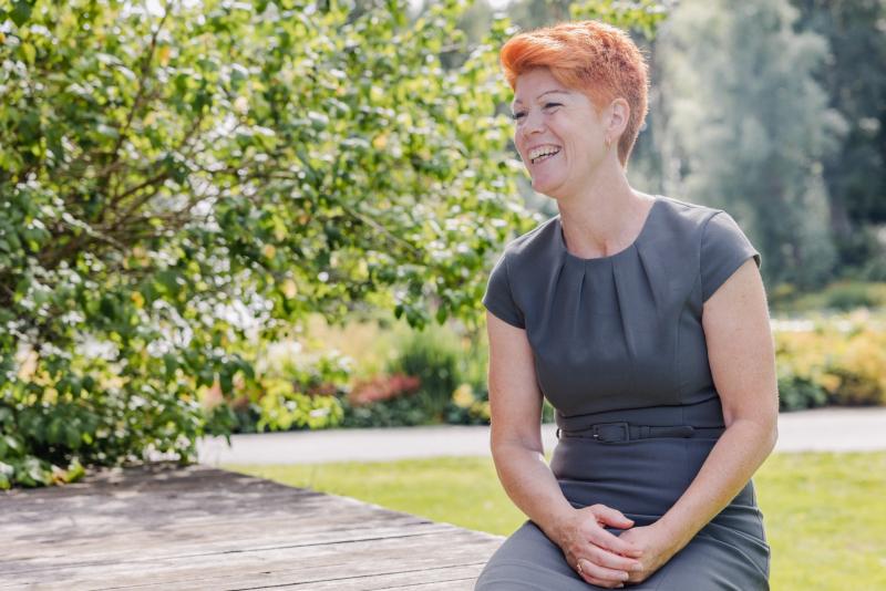 En person med kort rött hår och ljus skjorta med knappar står leende med armarna i kors framför en färgglad geometrisk bakgrund.