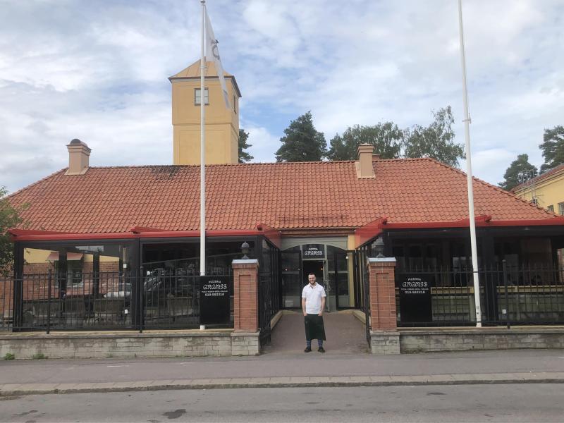 En person i vit skjorta står utanför en byggnad med rött tegeltak och torn. Två flaggstänger ramar in entrén.