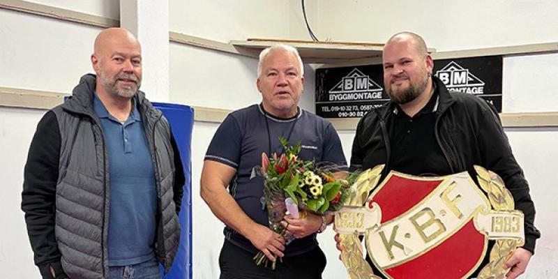 Tre män står inomhus; mannen i mitten håller i en blombukett, medan mannen till höger håller i en stor K.B.F.-sköld.