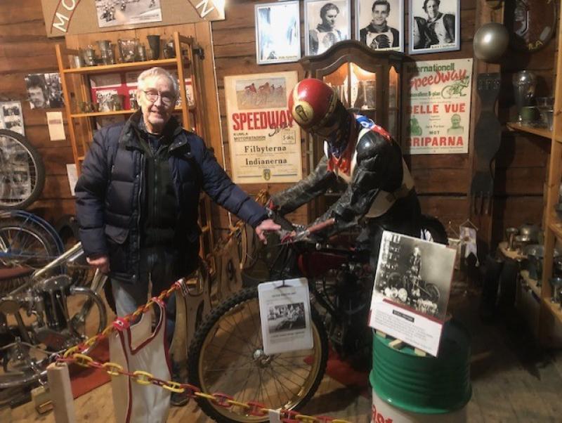 Man står bredvid en vintage motorcykelskärm med en skyltdocka i racingutrustning. Inramningen är ett museum med olika minnen och fotografier på träväggar.