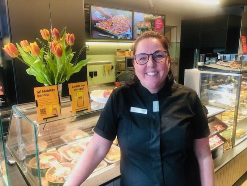 En kvinna i svart uniform står och ler bakom en glasdisk med bakverk på ett modernt café eller bageri.