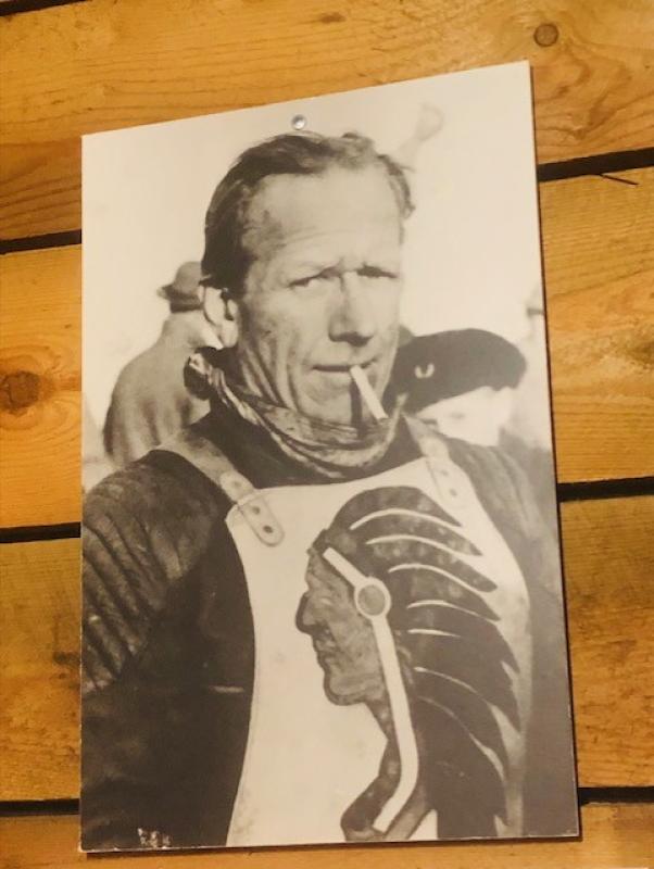 Svartvitt foto av en man med en cigarett, klädd i ett skyddsplagg med ett indiansk huvudbonads emblem, fäst på en trävägg.