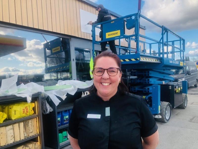 En kvinna i en svart skjorta ler mot kameran. Bakom henne arbetar en person på en blå upphöjd plattform nära en byggnad med stora fönster.