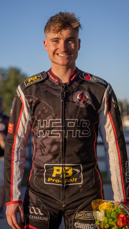 En person i en motorcykeldräkt och skyddsutrustning står utomhus och håller i en bukett blommor och ler direkt mot kameran.