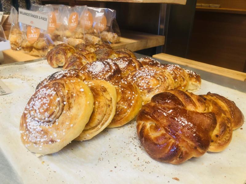 En bricka med nybakade bakverk, vissa toppade med pärlsocker, på pergamentpapper i en bagerimiljö. Förpackade bakverk syns i bakgrunden.