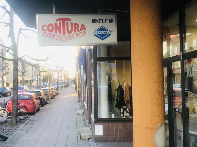 Gatuvy över Contura Konstlist ABs skyltfönster med skylt, klädesplagg och reflexer i fönstret. Trottoar, parkerade bilar och kala träd kantar gatan.