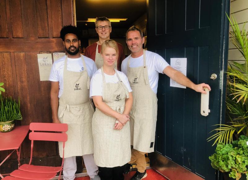 Fyra personer med rutiga förkläden står framför en dörröppning och poserar för ett gruppfoto. En röd stol och några växter syns i närheten.
