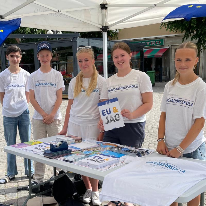 En grupp på fem ungdomar står bakom ett bord under ett tält, håller i material och bär 