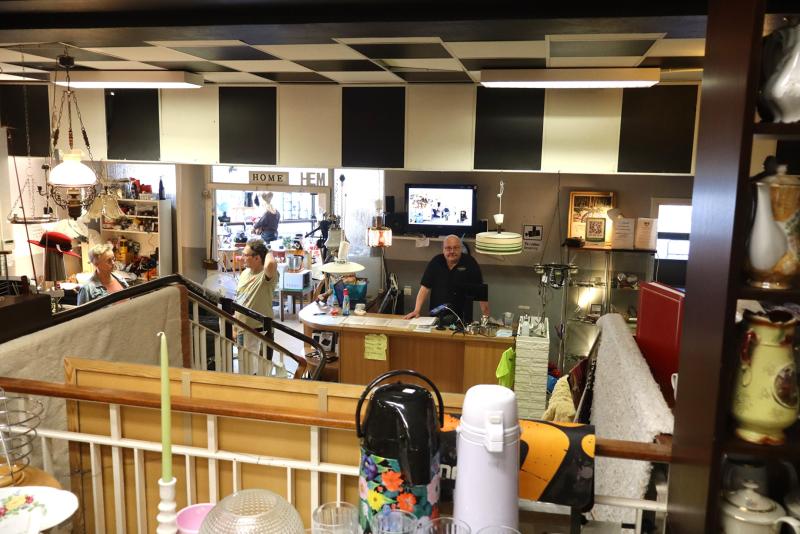 Interiör i en vintagebutik med en man bakom disken, olika föremål utställda och kunder som tittar runt i bakgrunden.