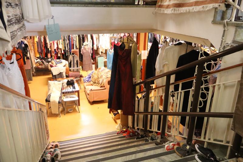 Utsikt från en trappa som leder ner till en secondhandbutik i källaren, med kläder på galgar, skor på trappstegen och möbler mitt i rummet.