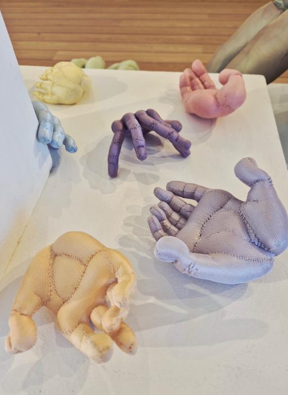 Flera handskulpturer i tyg i pastellfärger (gult, blått, lila och rosa) visas på en vit yta och ett trägolv.