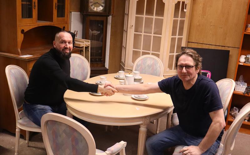 Två män sitter vid ett runt matbord i ett rum med trämöbler, skakar hand och är vända mot kameran. Det finns tekoppar och fat på bordet.