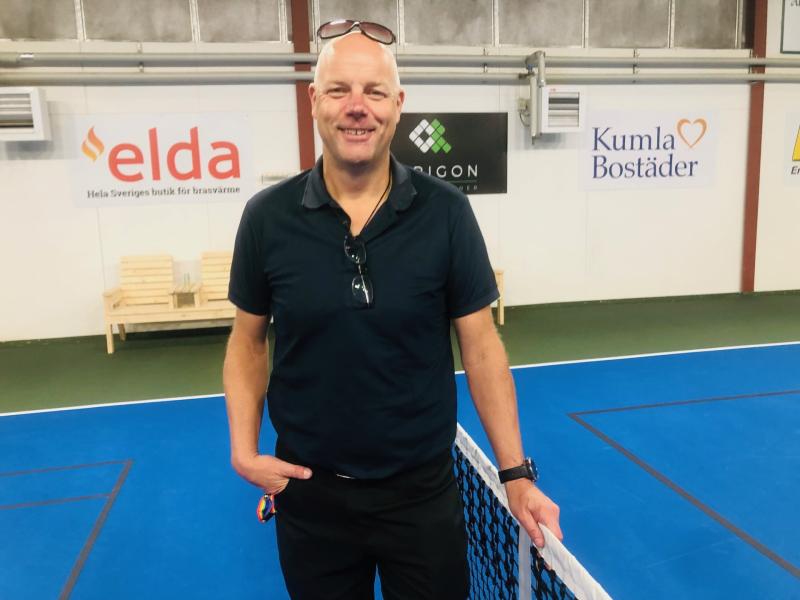 En man i svart skjorta och solglasögon står vid ett tennisnät på en inomhusbana med sponsorskyltar på väggen bakom honom.