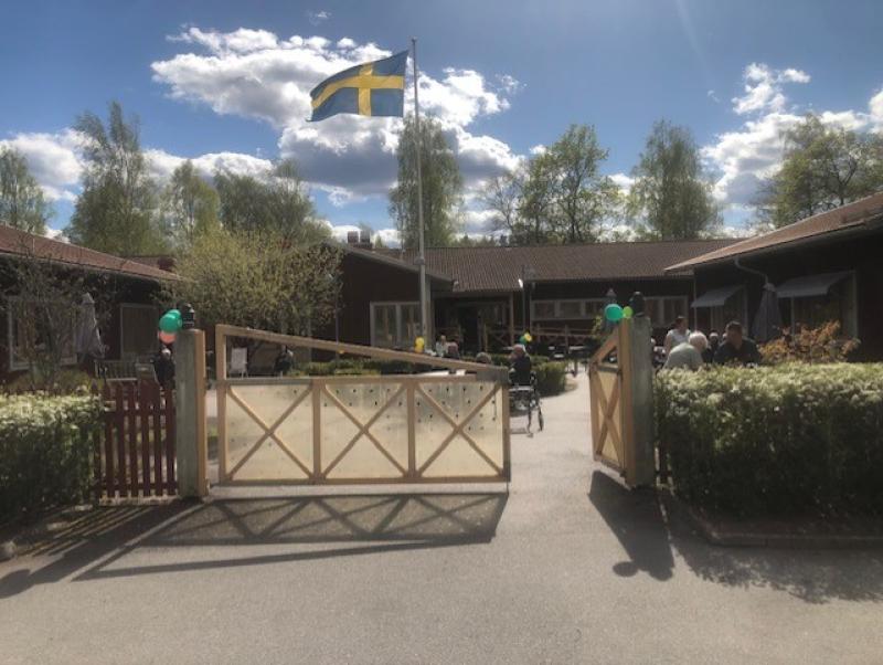En svensk flagga vajar ovanför en grindad entré till en byggnad med människor samlade utanför en solig dag, omgivna av grönska och dekorerade med ballonger.
