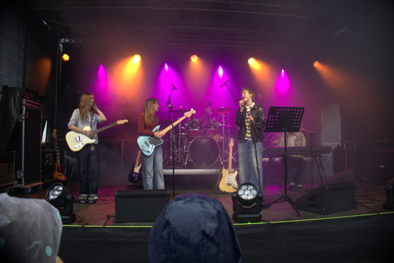 Ett band uppträder på en utomhusscen med färgglada lampor, med tre gitarrister och en sångare längst fram och en trummis i bakgrunden.