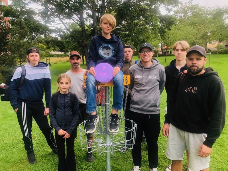 En grupp på åtta personer poserar utomhus runt en discgolfkorg; en pojke sitter på korgen och håller i en lila disc.