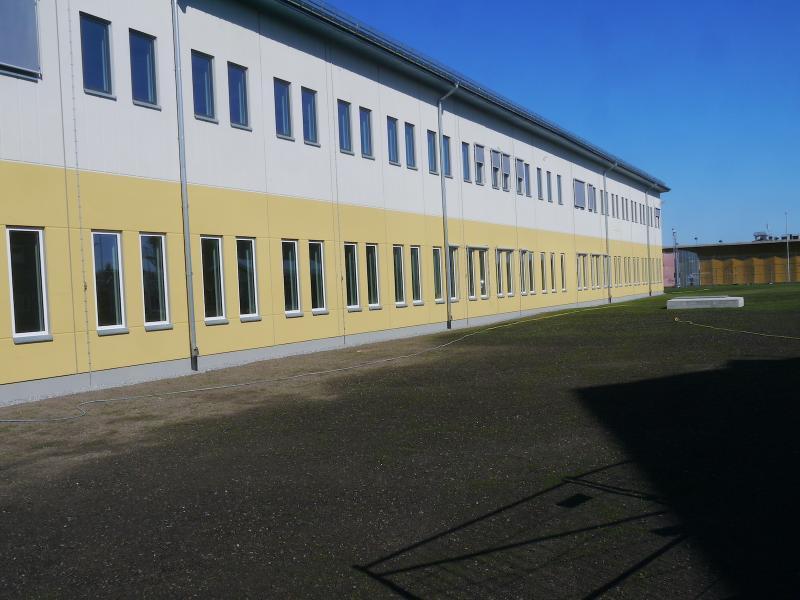 En tvåvåningsbyggnad med rader av rektangulära fönster på båda våningarna, gula och vita ytterväggar och en asfalterad gård framför under en klarblå himmel.