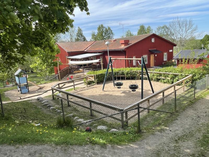 Inhägnad lekplats med gungor och lekställning, omgiven av träd och grönska, bredvid en röd byggnad på en solig dag.