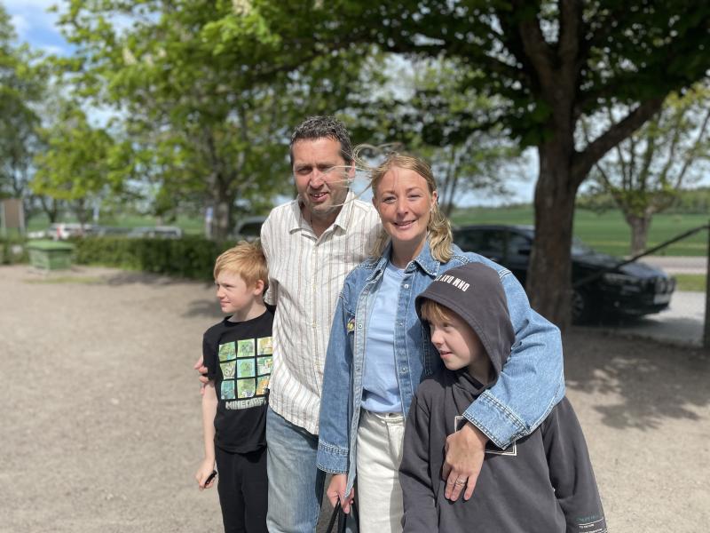 Fyra personer står utomhus en solig dag, med två vuxna som poserar tätt tillsammans och två barn som står framför dem. Träd och en parkerad bil syns i bakgrunden.