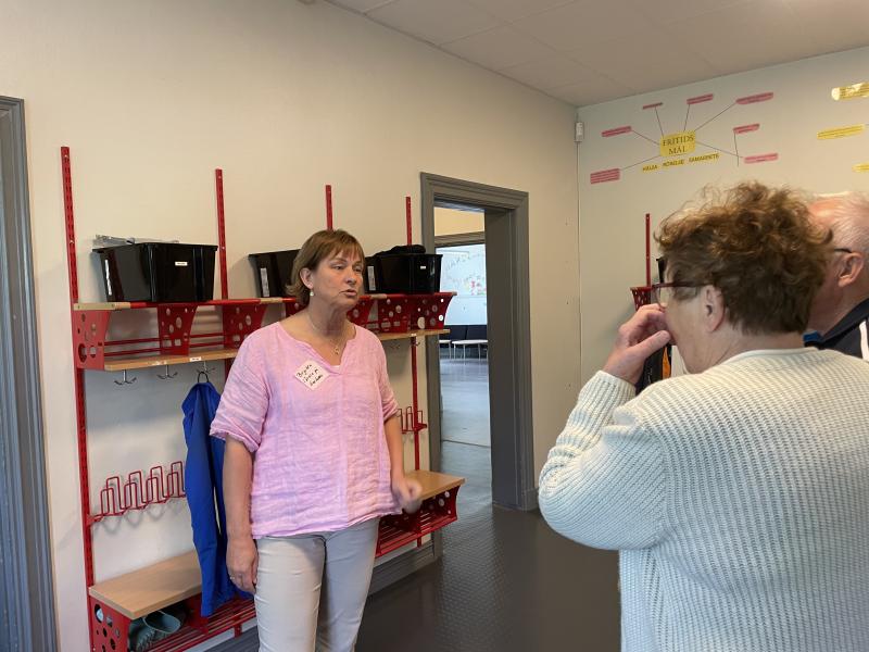 En kvinna i rosa skjorta pratar med två äldre vuxna i en hall med röda krokar, hyllor och svarta förvaringslådor på väggen.