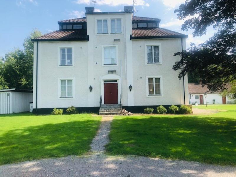 Ett stort, vitt trevåningshus med röd dörr, svarta lister och sluttande tak, beläget på en grön gräsmatta med en grusgång som leder till entrén.