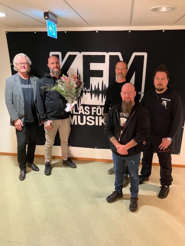 Fem män står inomhus framför en stor svart KFM-skylt; en håller i en blombukett och alla är vända mot kameran.