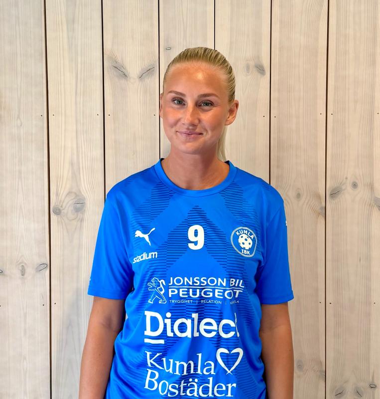 En kvinna klädd i en blå fotbollströja med nummer 9 står framför en ljus trävägg, tittar lite åt sidan och ler.