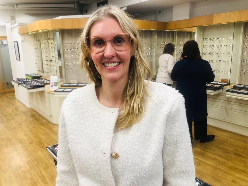 En kvinna med glasögon och en vit strukturerad rock står i en glasögonbutik och ler mot kameran. Två personer tittar på glasögonbågar i bakgrunden.