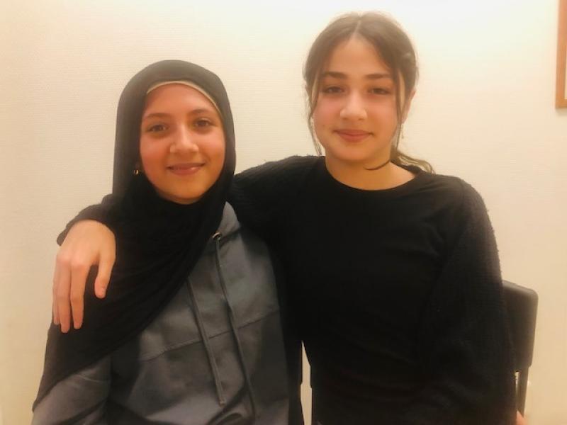 Två unga kvinnor sitter tätt intill varandra, den ena med armen runt den andra. Båda ler mot kameran; den ena bär hijab och den andra en svart tröja.