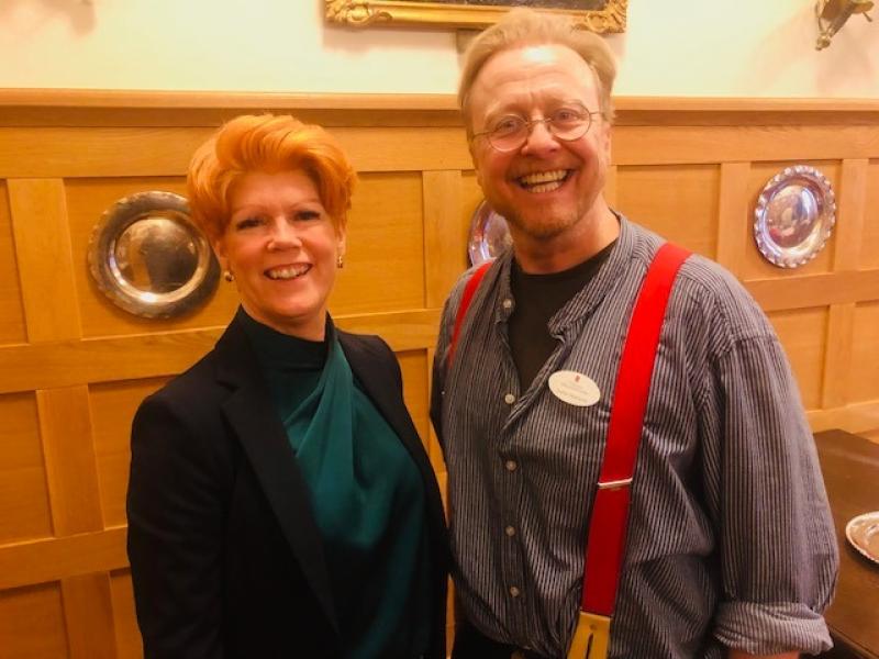 Två vuxna står sida vid sida och ler inomhus; den ena bär kavaj, den andra skjorta med röda hängslen och namnbricka. Träpanelvägg med silverplattor i bakgrunden.