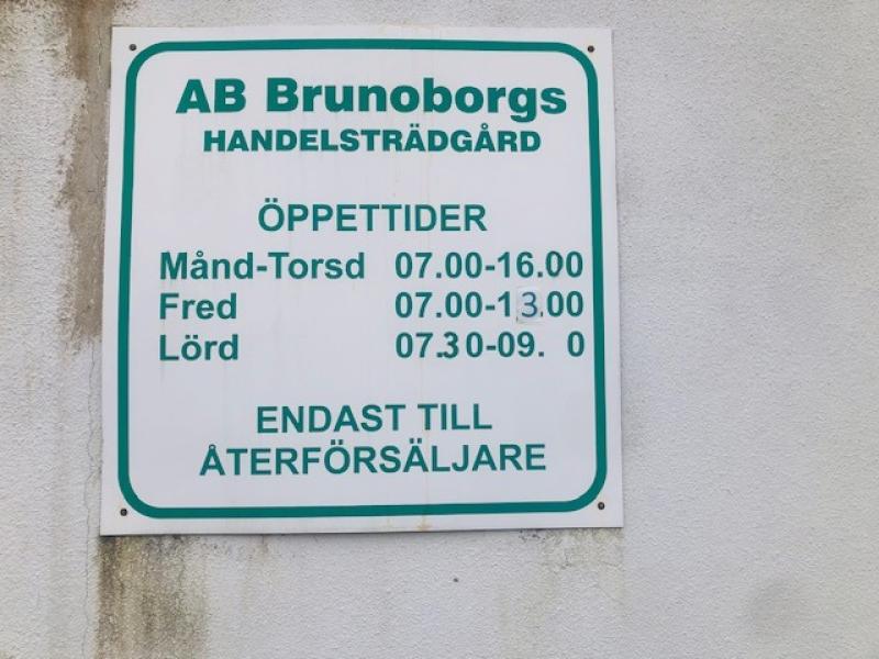 En vit skylt med grön text visar AB Brunoborgs Handelsträdgårds öppettider och anger att tillträde endast är för återförsäljare.