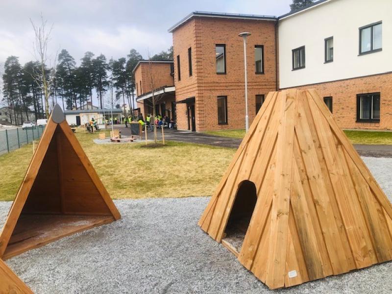 Lekplats utomhus med två tältliknande lekställningar i trä, gräs- och grusplan samt en skolbyggnad i tegel i bakgrunden.