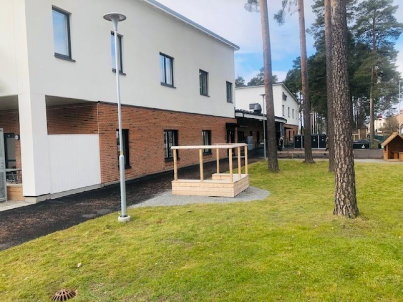 En tvåvåningsbyggnad med vit och tegelfärgad exteriör står nära en gräsyta med en liten träkonstruktion och höga träd i bakgrunden.